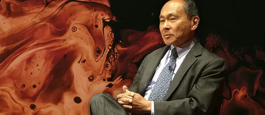 Man in a suit seated against an abstract background with earthy tones, appearing contemplative, possibly engaged in a discussion or interview. Man in a suit seated against an abstract background with earthy tones, appearing contemplative, possibly engaged in a discussion or interview.