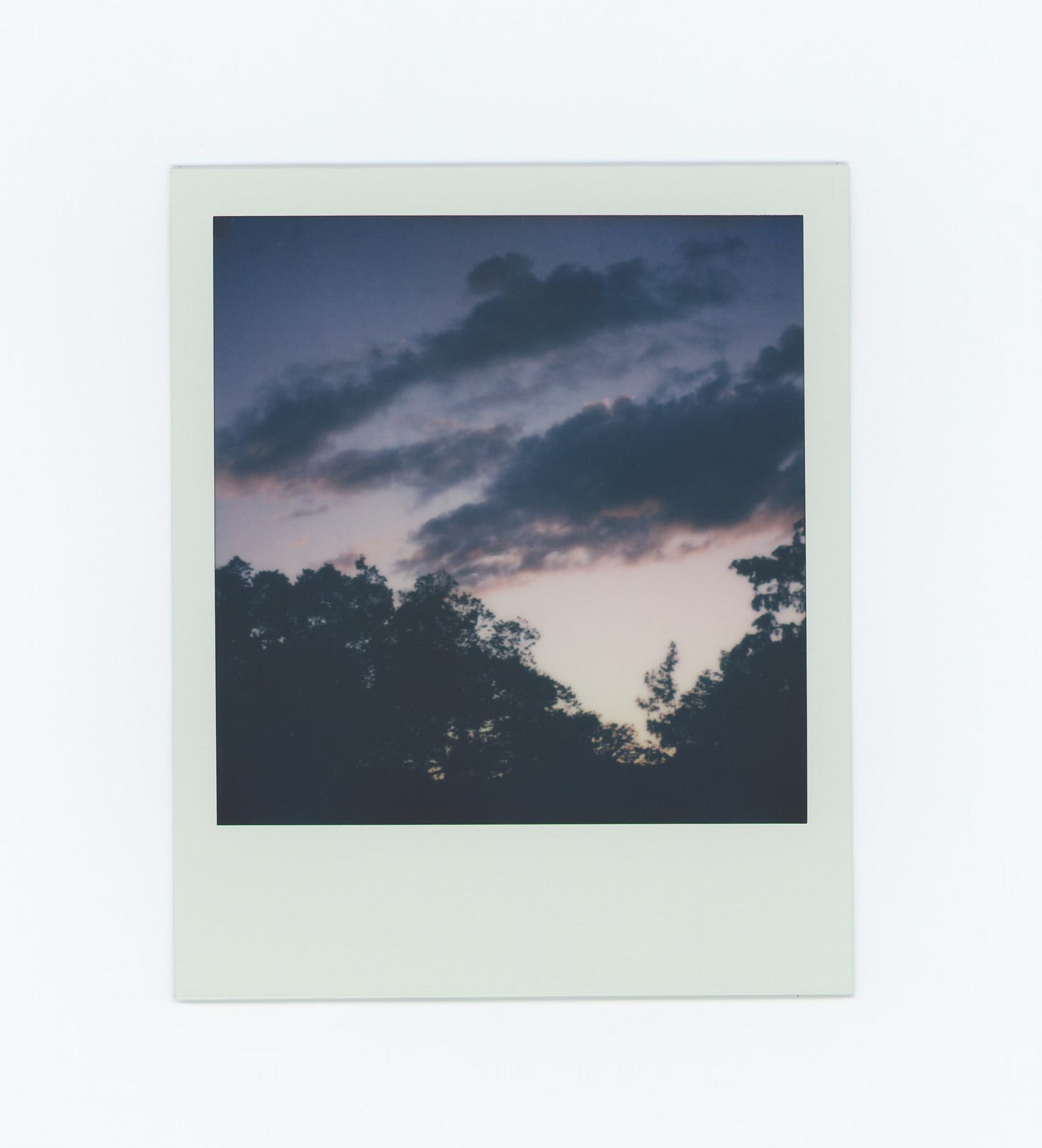A polaroid at sunset, blue sky fading to light pink, clouds dark blue/gray. Silhouette of trees along the bottom. 