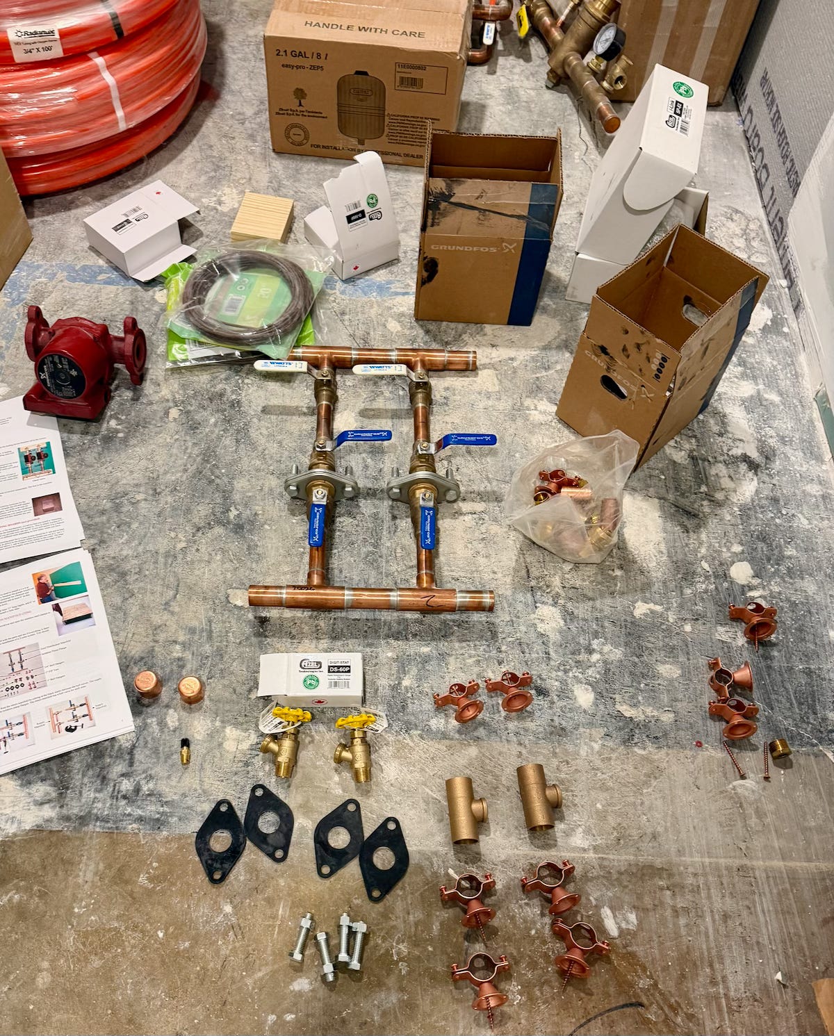 A variety of plumbing and radiant heating components are laid out on a basement concrete floor, including copper piping with blue-handled valves, fittings, pumps, and boxes of supplies. Additional parts such as clamps, gaskets, and instruction sheets are scattered around, with stacks of red PEX tubing visible in the background.
