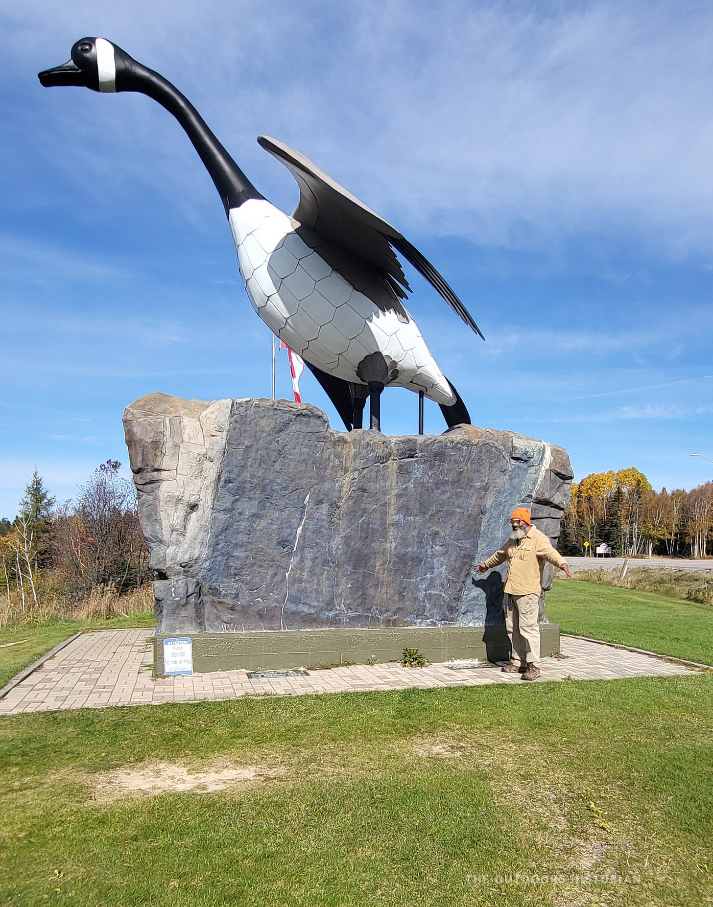 A photo of Eugene standing in front of the giant Wawa Goose statue on a sunny day. He has his arms stretched out, mimicking the goose's outstretched wings.