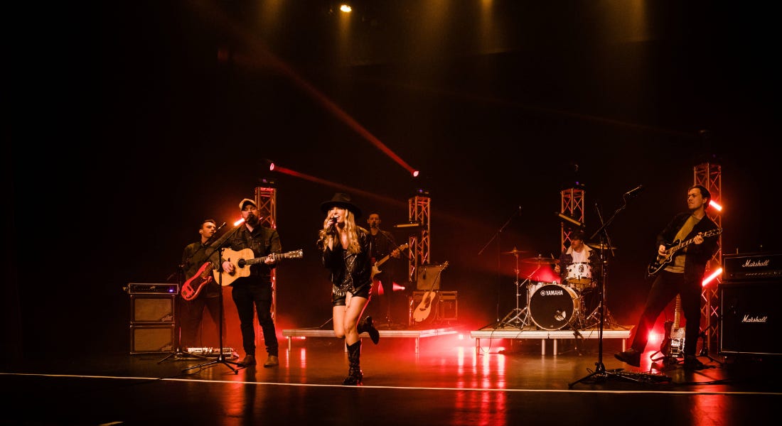 A stage with six people standing on it performing, with red lights around it and a black backdrop