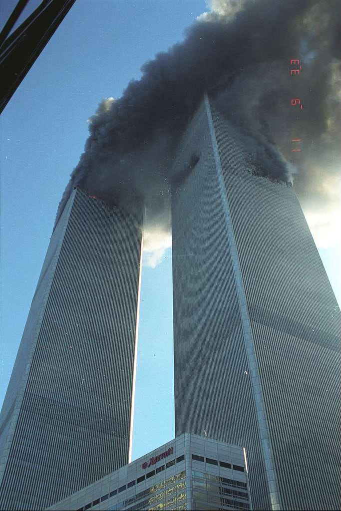 Both World Trade Center towers burn and emit thick black smoke into a clear blue sky on September 11, 2001, shortly after being struck by hijacked airliners. The Marriott Hotel is visible at the base.