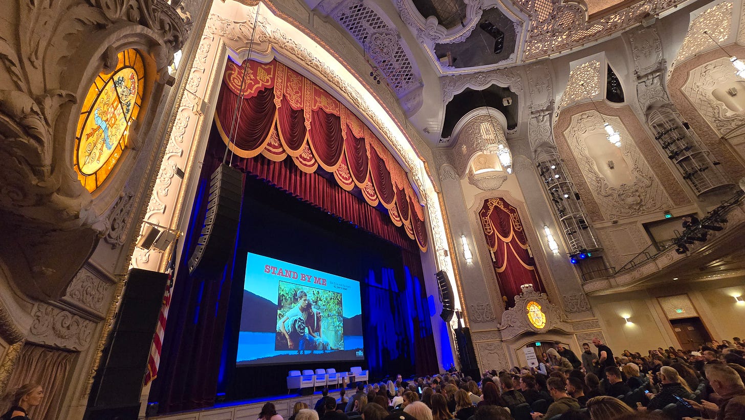 The inside of the theater, with stained glass and fancy sculptures, a big red stage curtain, and a big screen on the stage displaying the name of the movie, "Stand By Me"