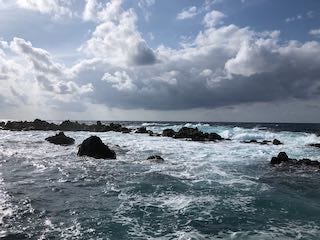 Photos of black rocks in a frothy ocean, a stone wall in front of fields and a village, a town plazaa with colorful buildings and orange tile roofs.