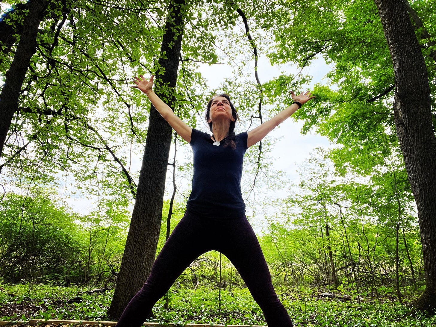 Woman standing in a wide-legged stance with arms reaching up to the sky in the middle of a forest