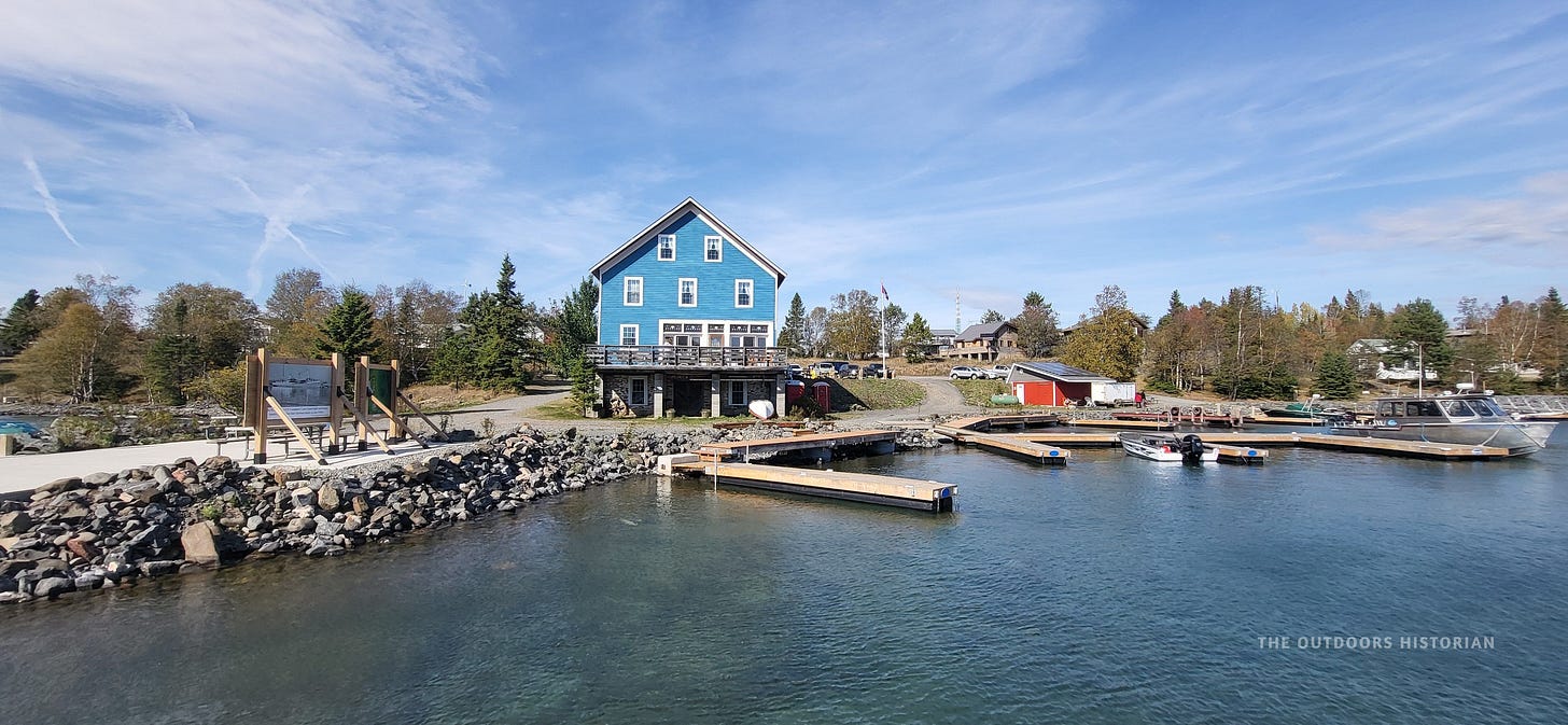 A historic two-story building with blue wood siding and white trim. A wooden dock sits in the foreground, and a Canadian flag flies from the porch.