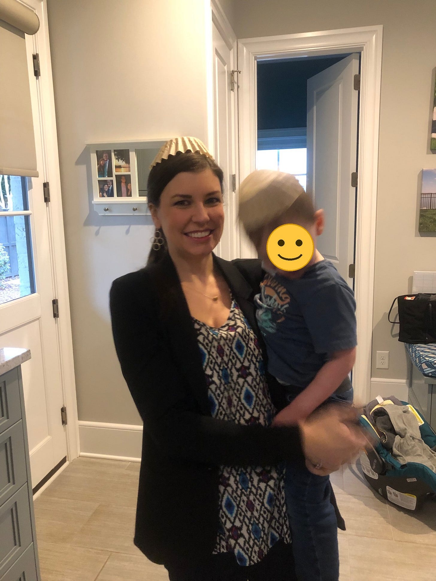 A woman in a blazer stands in a hallway holding her young child on her hip, a car seat on the floor behind her, ready to leave for work; a smiley emoji covers the child's face for privacy. A woman in a blazer stands in a hallway holding her young child on her hip, a car seat on the floor behind her, ready to leave for work; a smiley emoji covers the child's face for privacy.