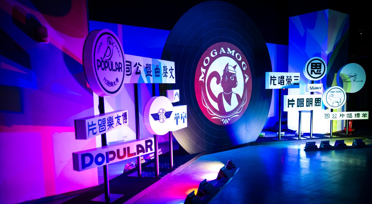 Black and white signs in multiple languages situated in front of a colorfully lit backdrop feature historic record label names