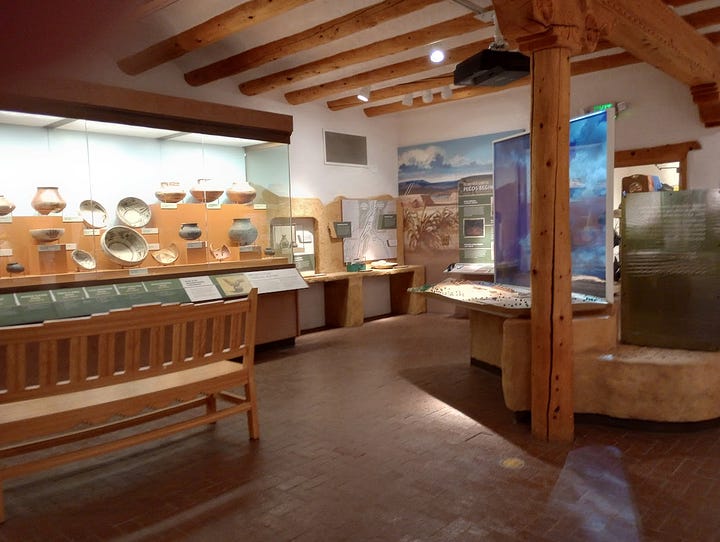Gift shop with books and souvenirs, museum displays including basketry and diorama of the Pecos Pueblo, view from cloister of semi-desert with trees
