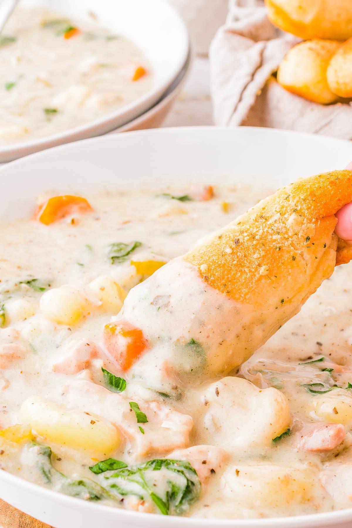 A bowl of creamy chicken gnocchi soup with visible chunks of chicken, carrots, and spinach. A hand dips a piece of bread into the soup. Another bowl and bread are blurred in the background. A bowl of creamy chicken gnocchi soup with visible chunks of chicken, carrots, and spinach. A hand dips a piece of bread into the soup. Another bowl and bread are blurred in the background.