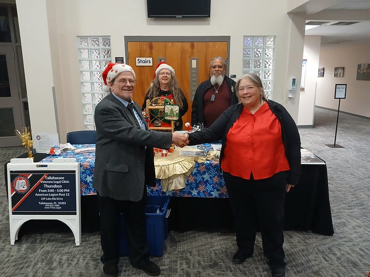 Two men stand on a stage under blue lighting. On the left, an older man in a gray blazer, tie, glasses, and a Santa hat gestures with a pointed finger. On the right, a bearded man in a bright blue shirt holds a folder with printed pages and looks at his hand as if responding. A microphone on a stand is beside them, and a music stand is in the foreground, suggesting a performance or reading. Also, A festive scene in a building lobby: four adults stand behind and around a holidayâdecorated information table. In front, a man in a Santa hat and a woman in a red blouse shake hands and smile. Behind them, two others (one wearing a Santa hat, another with a long white beard and dog tags) stand by the table, which is covered with a blue, starâspangled cloth, gold runner, ornaments, and brochures. A sandwichâboard sign on the left reads âTallahassee Veterans Legal Clinicâ with details for Thursdays at American Legion Post 13, 229 Lake Ella Drive, Tallahassee, Florida. The background shows a wooden door marked âStairs,â glassâblock windows, and a wallâmounted TV.
