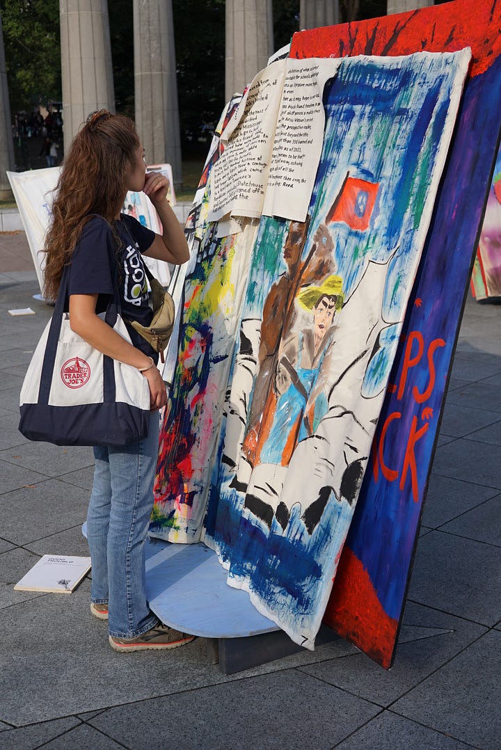 A woman holding a white bag examines a brightly colored painted giant book. A child uses a marker to add to a book made out of pillows and furniture. A group of people examines a colorful giant book with painted text and images. A giant, brightly colored book stands in front of a building with columns.
