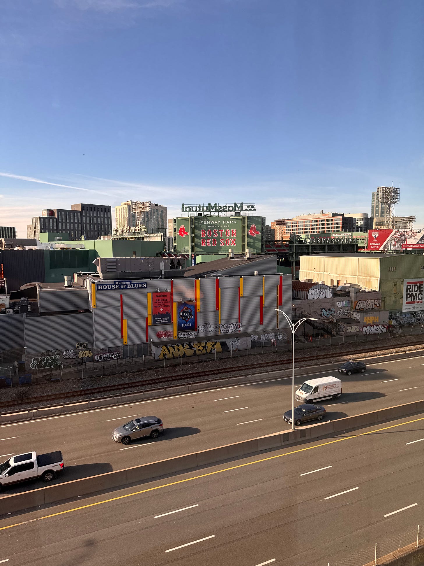 View of Fenway Park as seen from hotel window. The famous "green monster" wall stretches across the photo, with "Fenway Park: BOSTON RED SOX" in the foreground. Also in the foreground are the lanes of the Mass Pike with several passing cars. Blue sky in the background.