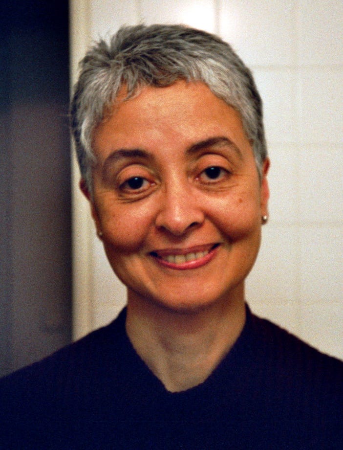 Photo of a woman smiling. She has cropped gray hair, gold earrings, dark eyes. She wears a black top and is against a white wall and ⅓ shadow on the left side.