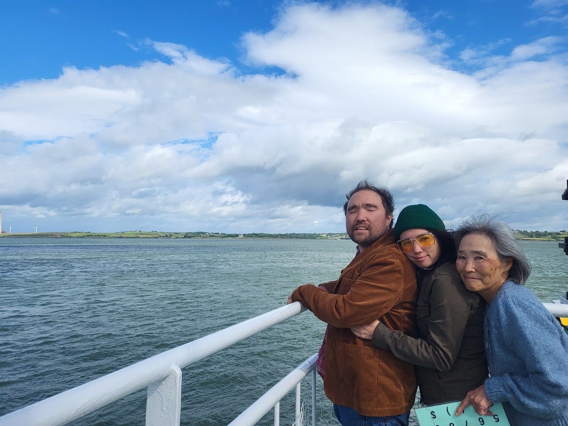 Danny, his sister, and his mom stand together on a ferry. Danny is leaning against the railing, overlooking the water. His sister is hugging him from behind, and their mother is hugging both of them from behind.