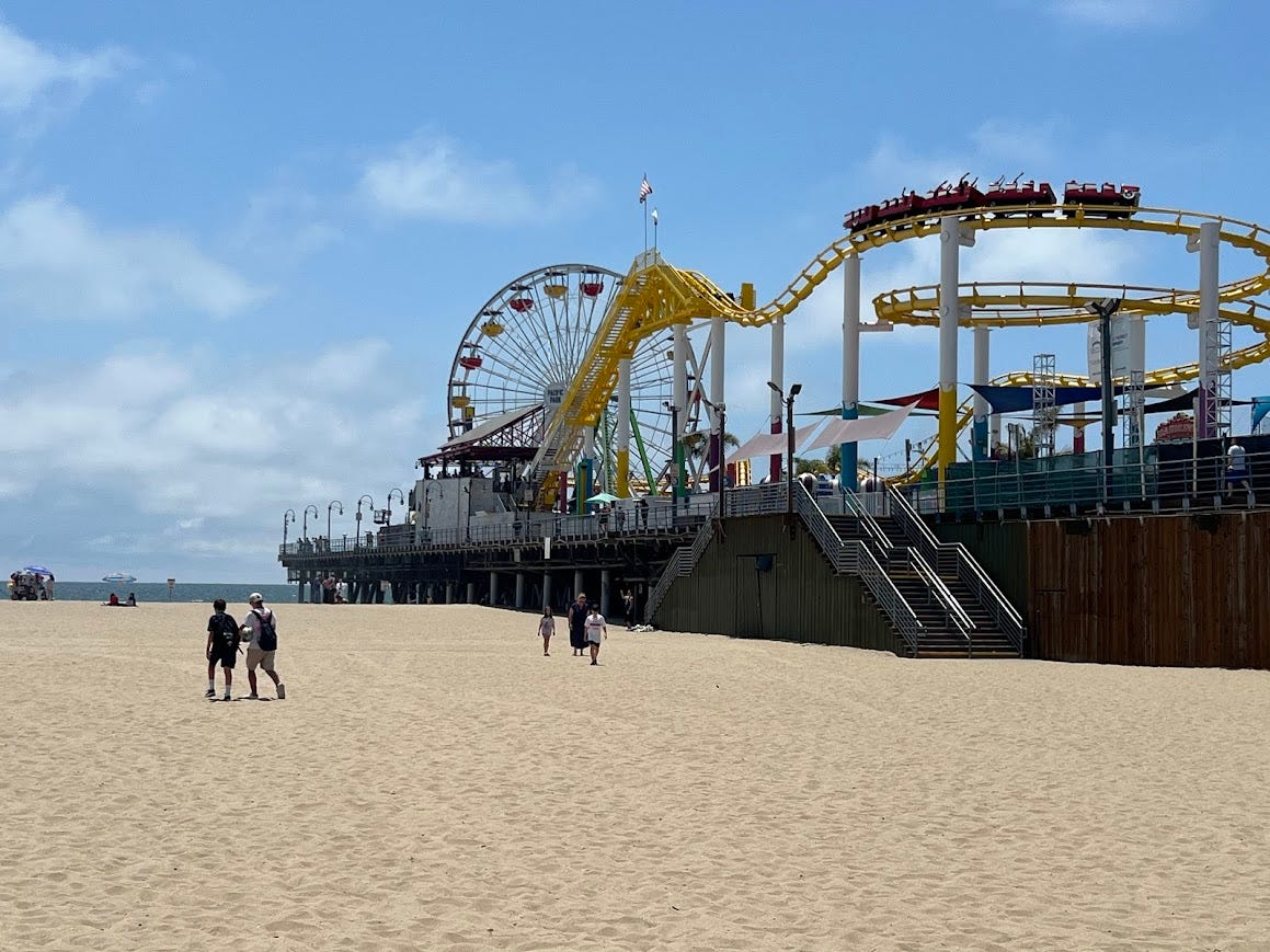 Wide photo of the Santa Monica pier showing a Ferris wheel, beach, and a rollercoaster.