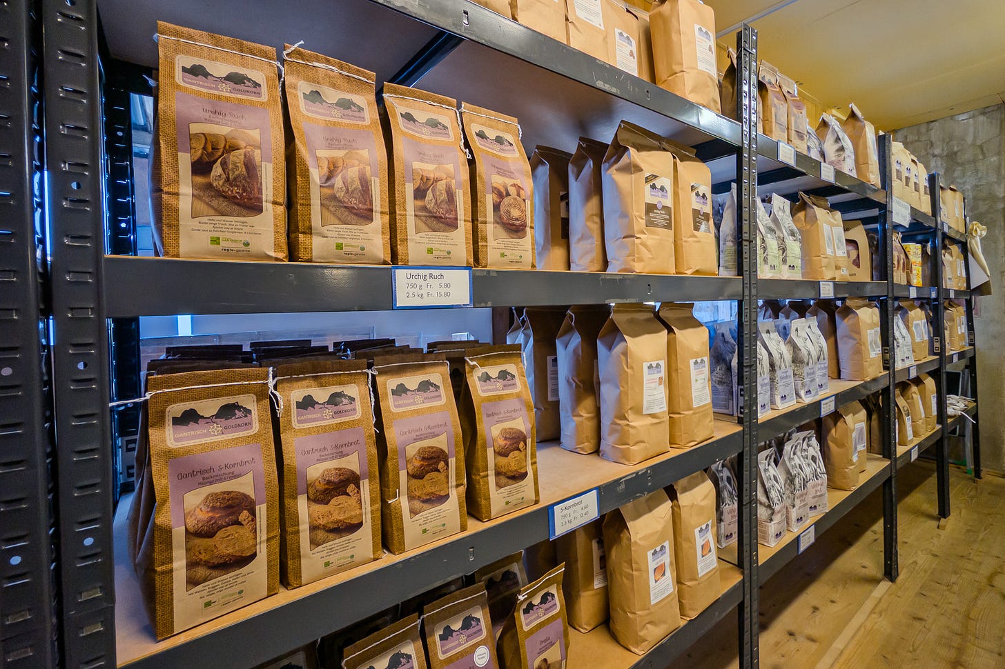 Im Verkaufsraum der Dittligmühle ziehen sich mehrere hohe Metallregale mit Holzfächern in die Tiefe des Raums. Darin stehen zahlreiche Papiersäcke mit Mehl, Brot- und Backmischungen, sauber geordnet und mit kleinen Preisschildern versehen. Der kleine Mühlenladen wirkt schlicht und funktional. Im Verkaufsraum der Dittligmühle ziehen sich mehrere hohe Metallregale mit Holzfächern in die Tiefe des Raums. Darin stehen zahlreiche Papiersäcke mit Mehl, Brot- und Backmischungen, sauber geordnet und mit kleinen Preisschildern versehen. Der kleine Mühlenladen wirkt schlicht und funktional.