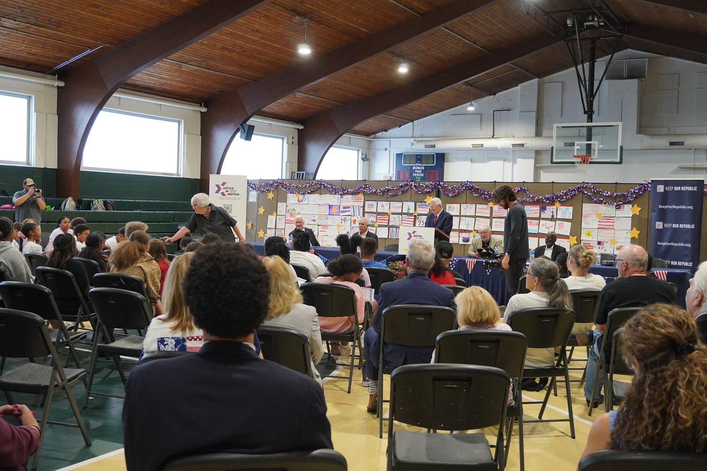 in a large recreational space with folding chairs, a group of judges presents at the front of the room infront of a bulletin board covered with cards in a large recreational space with folding chairs, a group of judges presents at the front of the room infront of a bulletin board covered with cards