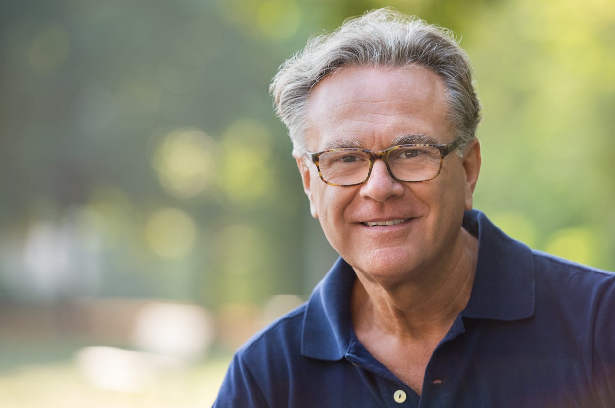 Smiling older man with gray hair and glasses in the outdoors. Blirry green foliage behind him.
