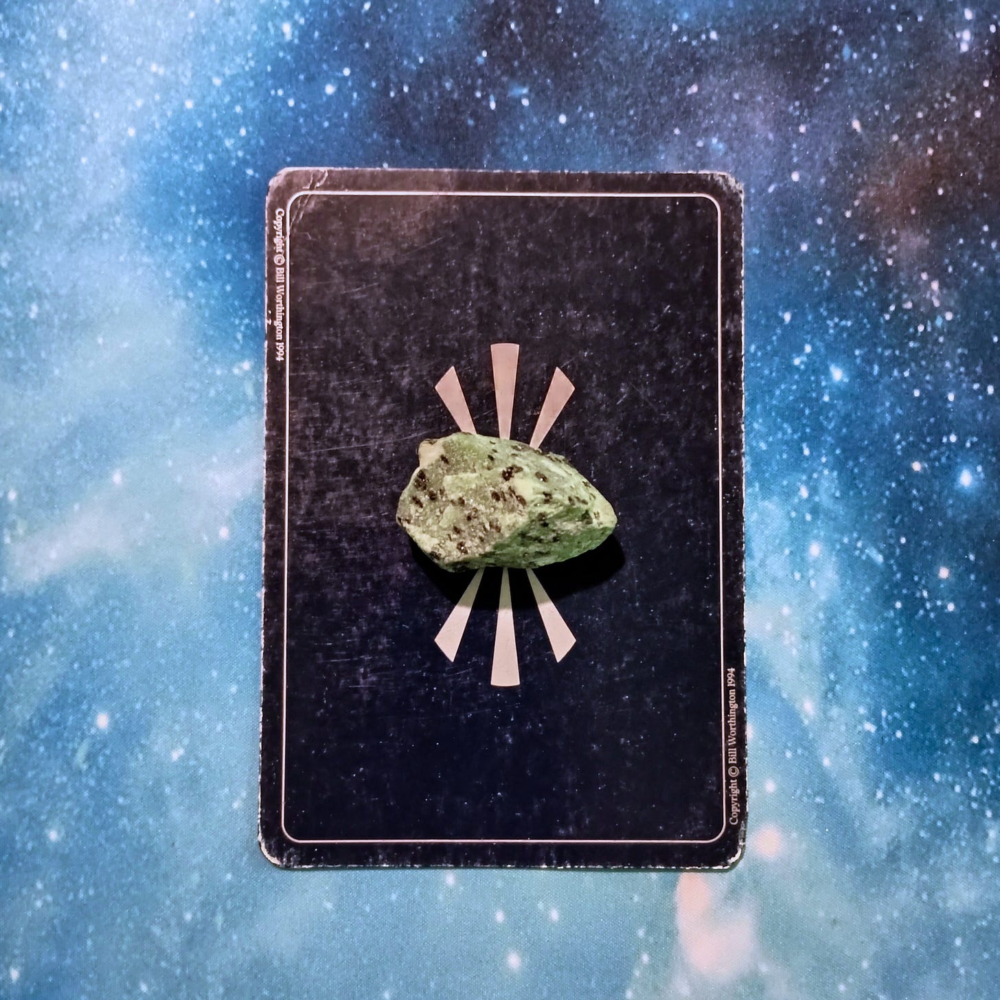 New Moon Magic: Your July 2025 Pick-a-Card Tarot Reading, pile 2 - ruby zoisite stone (green with black speckles) and an oracle card, face down, on a starry blue background