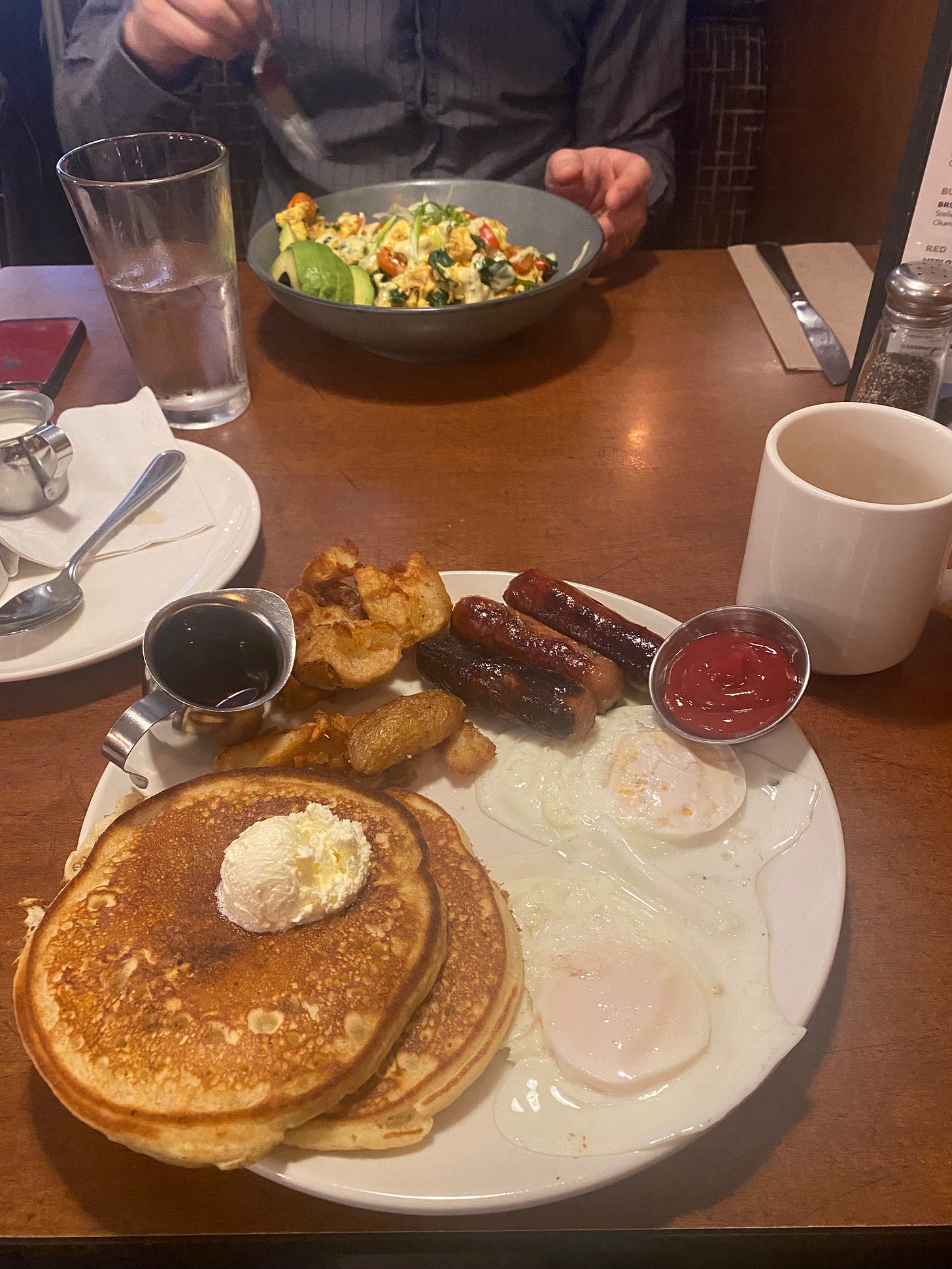 A big plate of diner breakfast: three sausages, a ramekin of ketchup, two over easy eggs, two pancakes with a scoop of whipped butter, smashed potato hash browns, and a little metal jug of syrup. Next to the plate is a white mug of coffee, and across the table Jeff has his fork poised over a bowl of scrambled egg and vegetable hash with avocado.