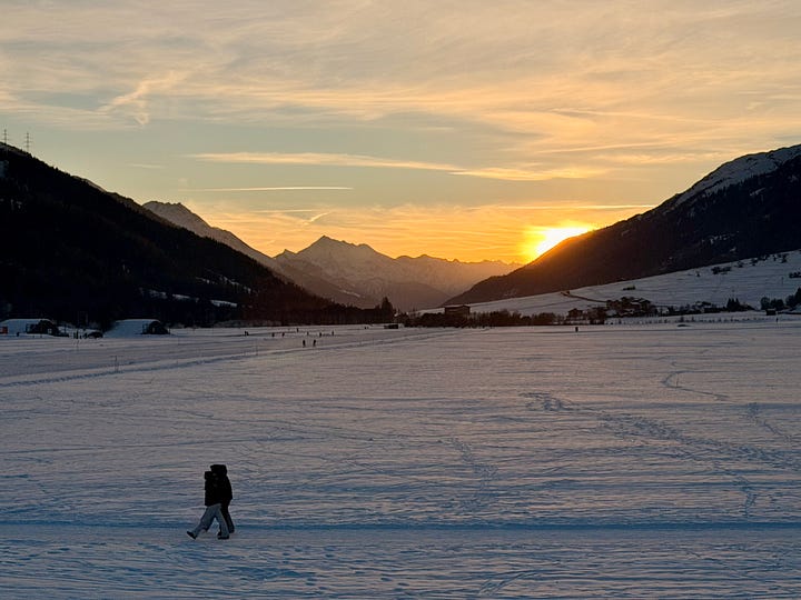 Left image, A snow-covered alpine valley plateau at sunrise. The peak of the centered Weisshorn mountain is sun kissed, and the sky around it tinted pink. The snow of the valley, still in shadows has a bluish tint; right image, the sun sets behind a mountain slope as view from a snow-covered valley plateau. Two walkers make their way along a groomed path in the foreground. In the background, the sharp, snow-covered Weisshorn mountain. The setting sun flashes a golden aura and tints the clouds above the mountains and snow on the valley floor orange.