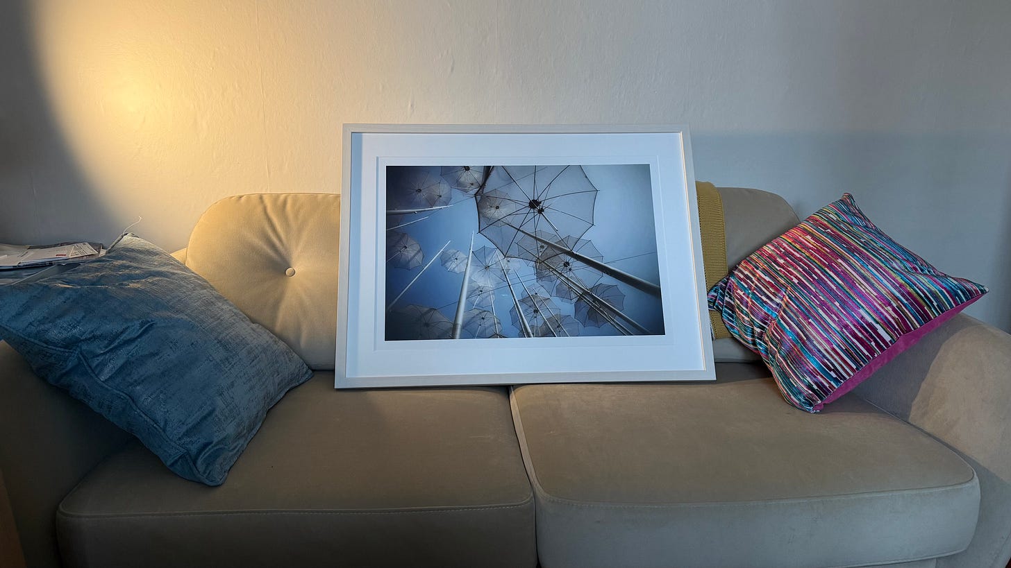 A framed photograph of the Thessaloniki Umbrellas sculpture rests on a cream sofa between a blue cushion and a colourful striped one. Soft daylight falls across the scene, highlighting the print’s pale tones and white frame.