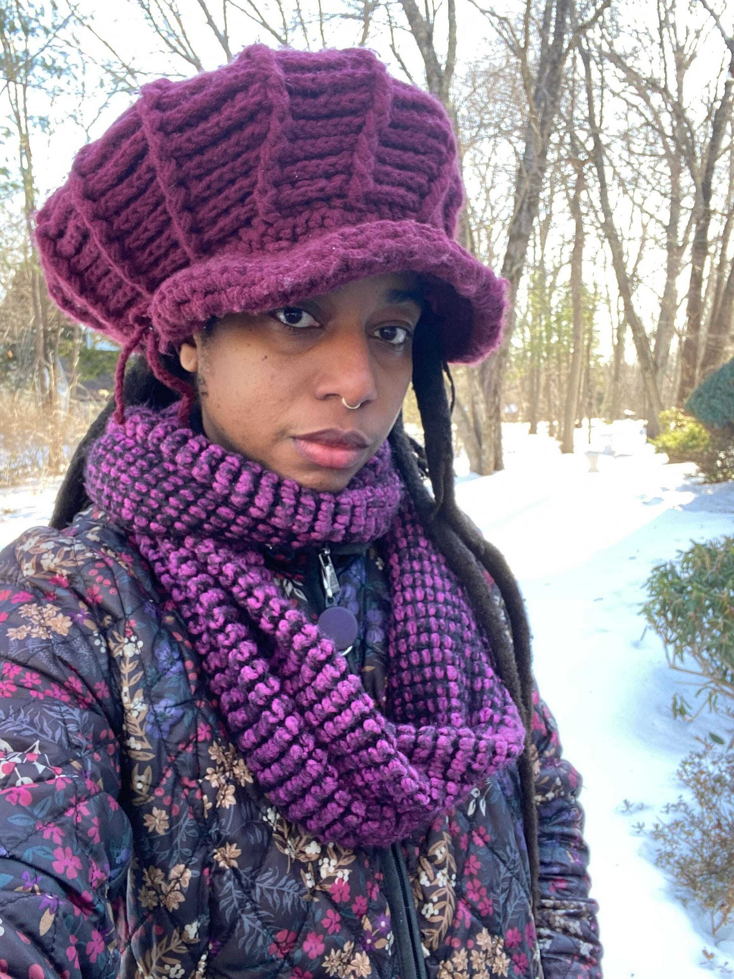 beautiful black woman with freeform locs standing in a winter landscape with snow. She stands out from the backdrop, wearing a large burgundy crochet hat, bright purple scarf, and a jacket with gold, black and lavender flower prints. beautiful black woman with freeform locs standing in a winter landscape with snow. She stands out from the backdrop, wearing a large burgundy crochet hat, bright purple scarf, and a jacket with gold, black and lavender flower prints.