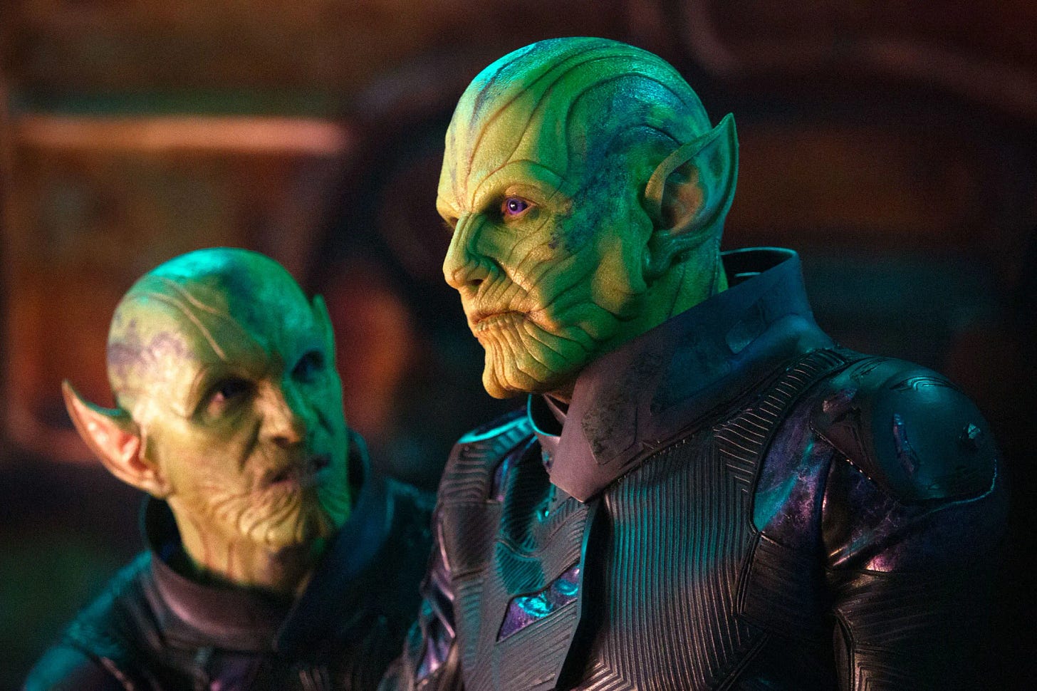 two green bald wrinkly skrulls, one looking at the other, the other looking offstage; they're both from chest up more or less and wearing form fitting tight futuristic clothes