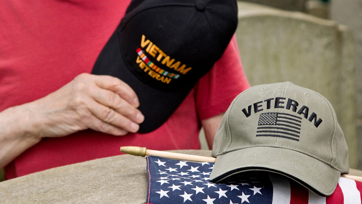 Man in red t-shirt, black hat in hand over heart. Black hat has Vietnam Veteran and ribbons on the front of the cap. Man leans over casket with small American and tan cap with Veteran and monotone black American flag on the front of the cap.