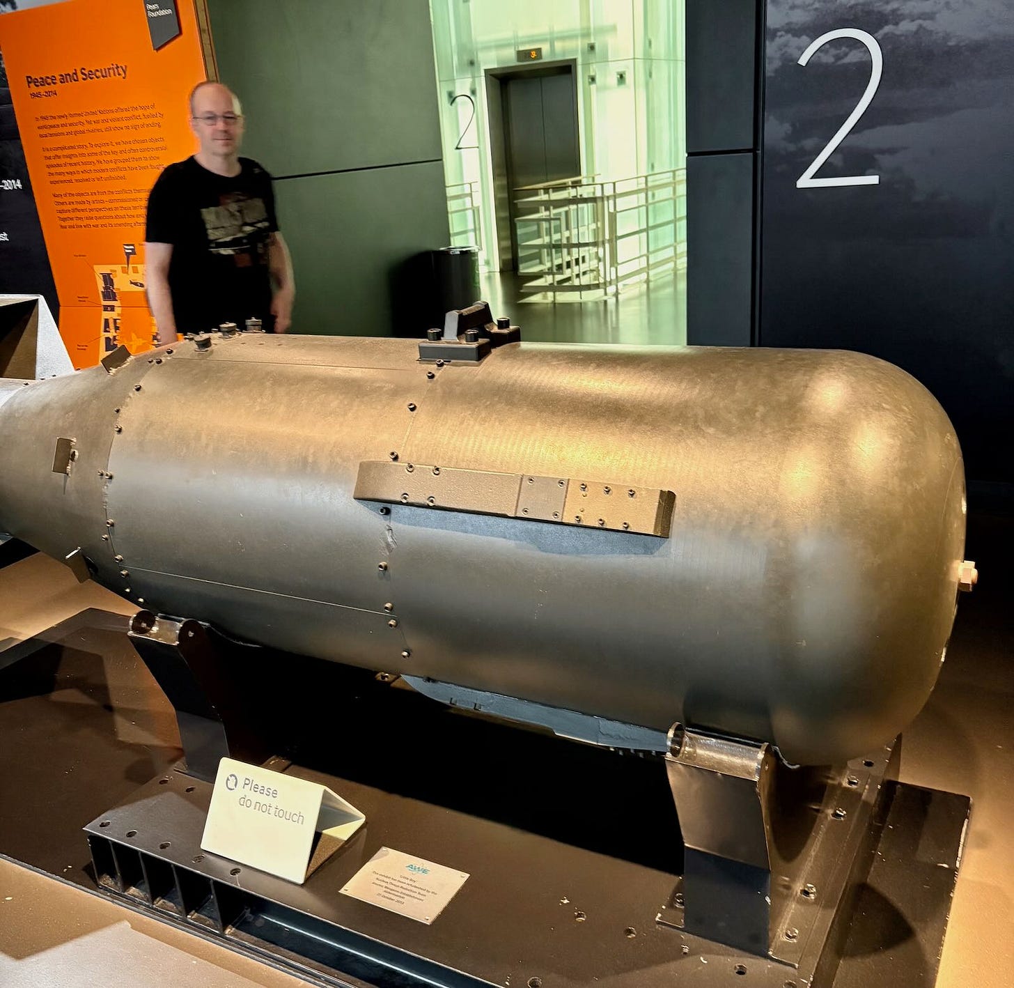 A man standing near the "Little Boy" atomic bomb replica at the Imperial War Museum London, showing its cylindrical shape.