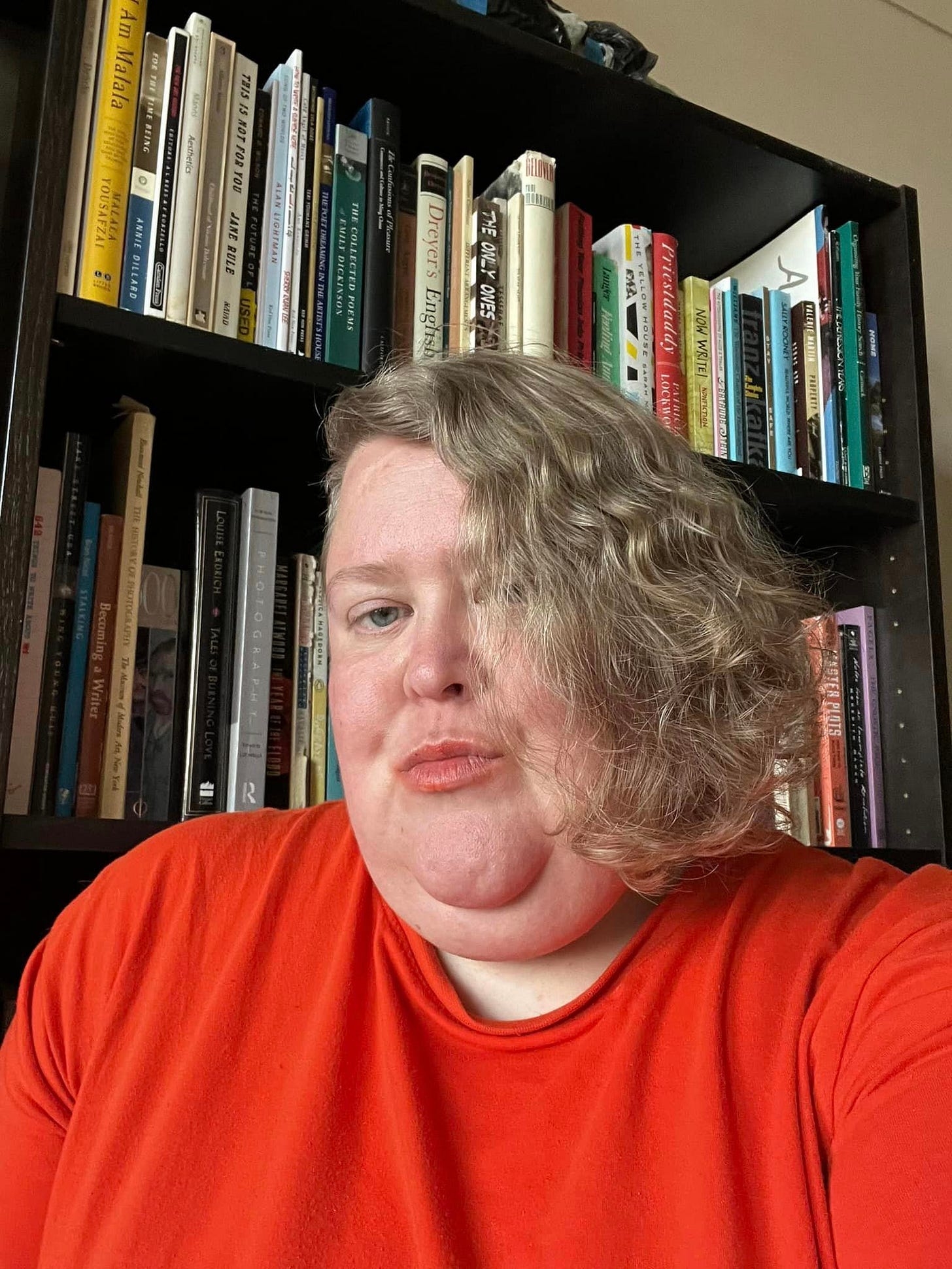 me, Alison, in front of my bookshelf looking different directions. My hair is blonde and almost shoulder length on one side. Shaved short on the other. I’m back to orange lipstick but not bothering with foundation