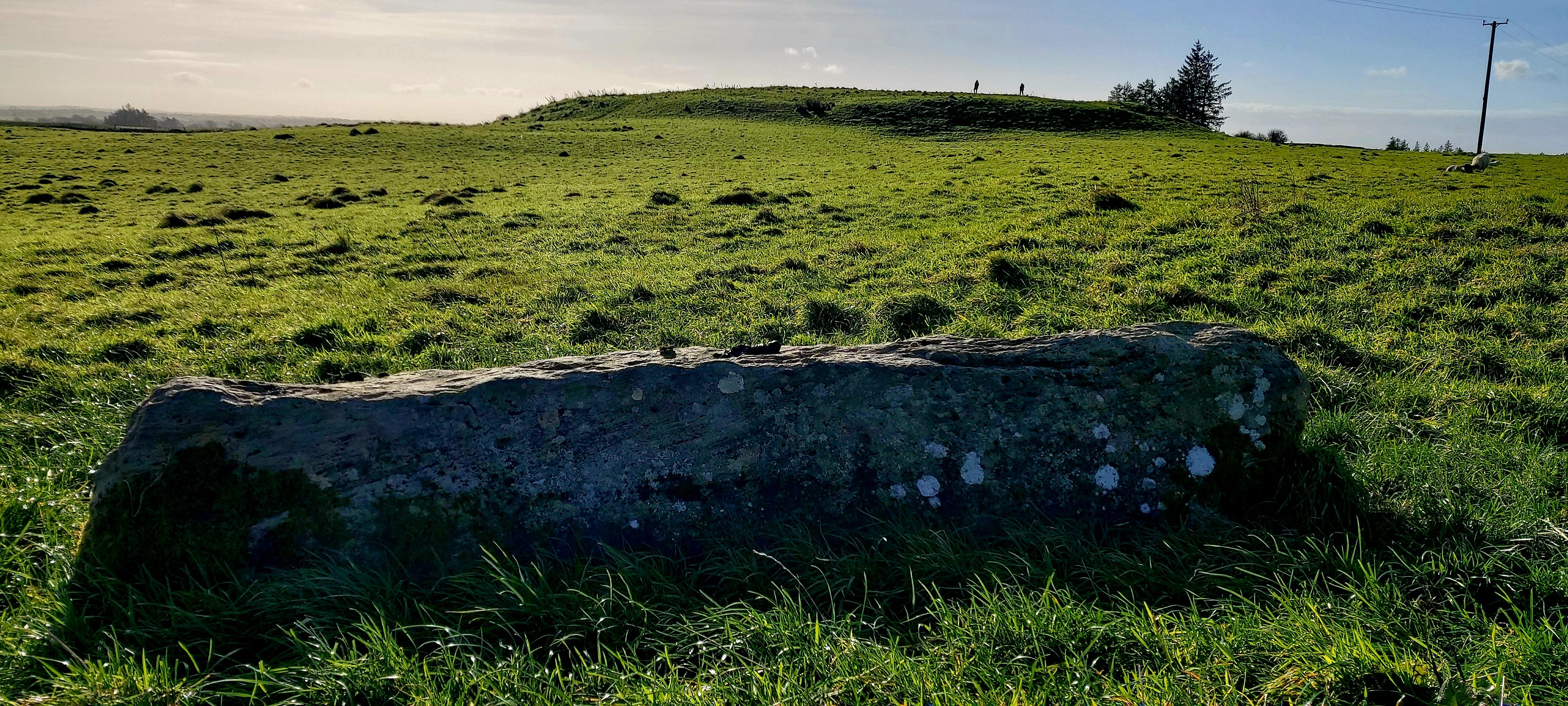 In the foreground of this image is a large flat recumbent standing stone; beyond it, the grassland rises into an enormous flat-topped mound, with two trees to the right. Two figures are walking along the top of the mound. In the foreground of this image is a large flat recumbent standing stone; beyond it, the grassland rises into an enormous flat-topped mound, with two trees to the right. Two figures are walking along the top of the mound.
