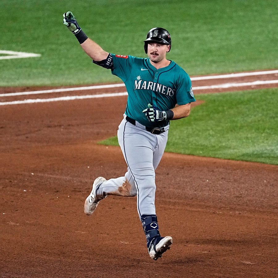 A baseball player wearing a green Seattle Mariners jersey with number 29 and matching cap runs on the green field with arms raised in celebration holding a bat in his right hand green batting gloves on both hands gray pants with green accents white cleats and black undershirt visible under jersey stadium lights and foul lines in background.