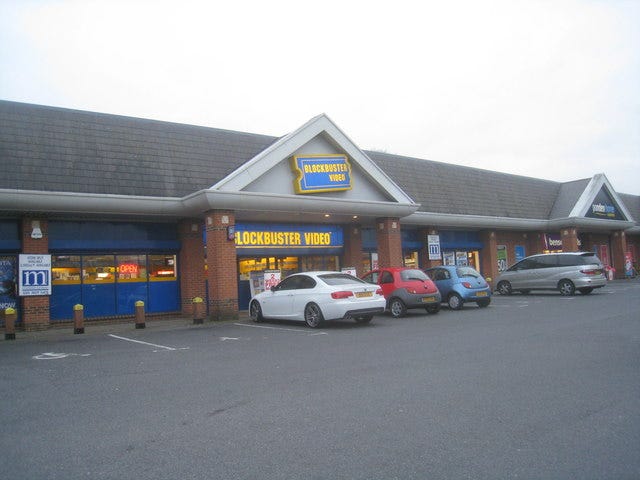 Blockbuster Video store in a shopping part, lit up from the inside, with cars parked outside.