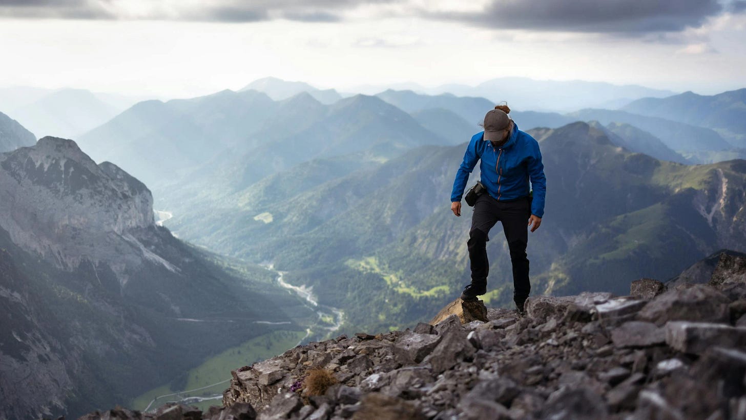 Hiker in blue jacket standing on rocky mountain summit, looking down at a breathtaking panorama of mountain ranges and valleys stretching into the distance under a cloudy sky.