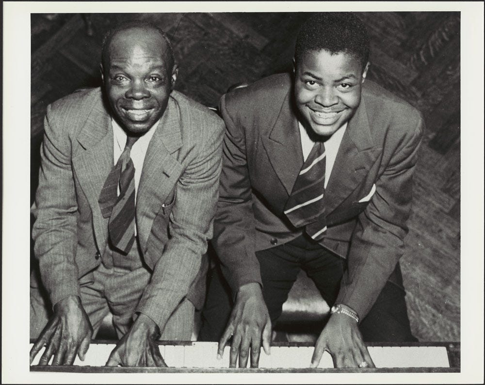 Black and white photograph of two black men - one older, and one in his late teens- both dressed in suits sitting at a piano looking up at the camera as their hands are placed side by side on the white ivory piano keys.
