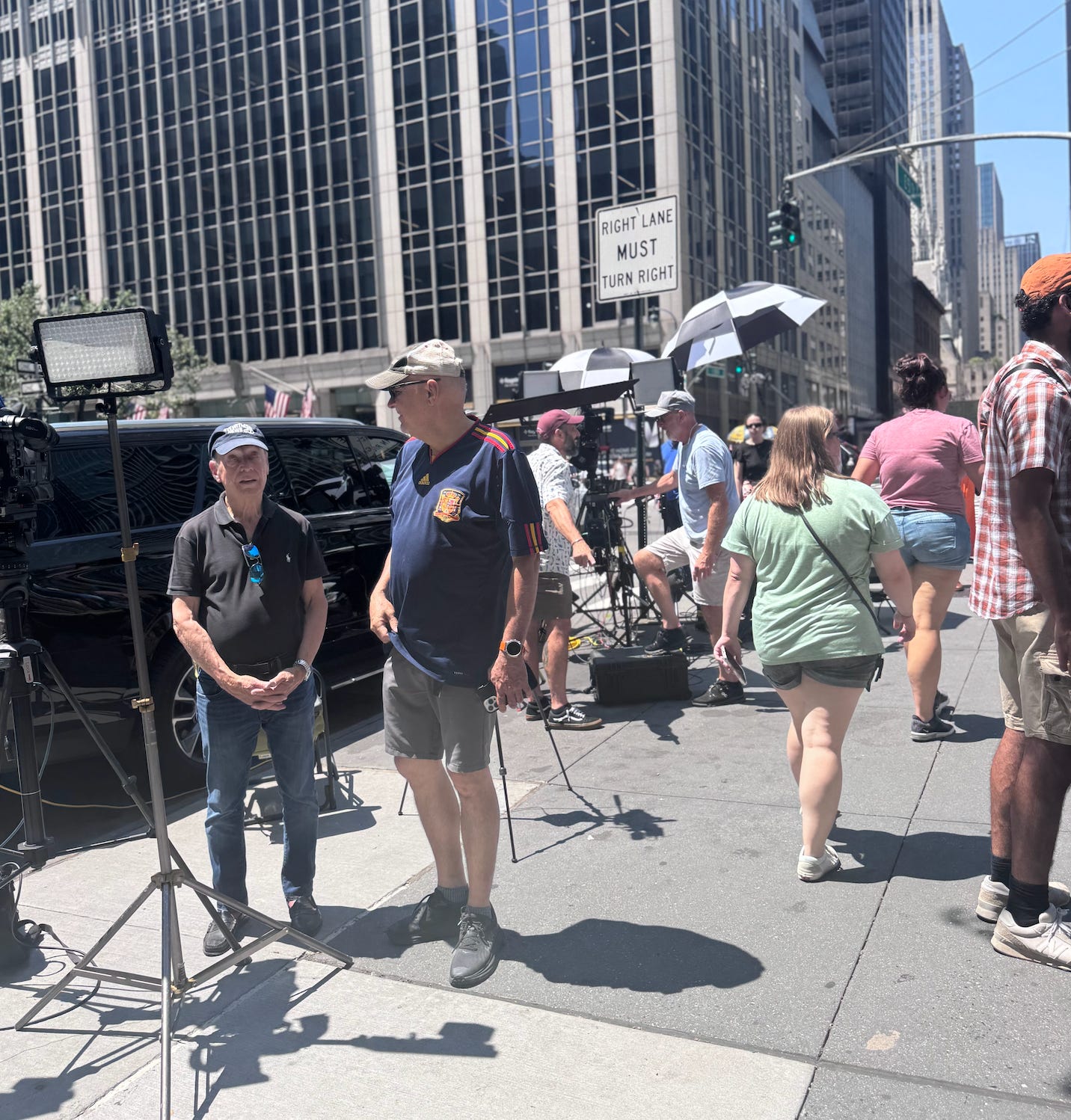 News teams are set up with lights and sun umbrellas on the sidewalk.