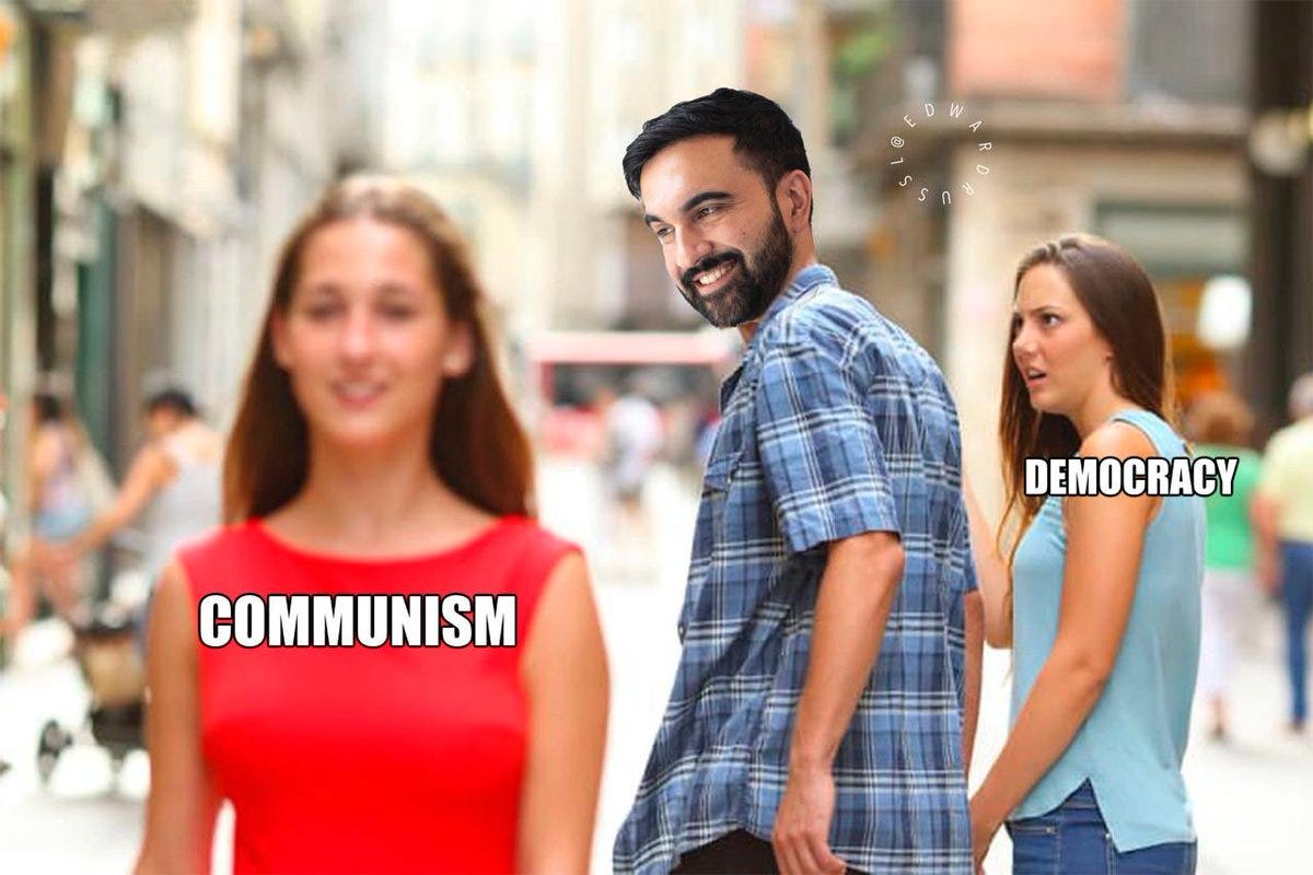 A man with short dark hair and a beard wearing a short-sleeved blue plaid shirt stands on a sunny urban street lined with buildings and pedestrians. He holds the hand of a woman on his left with long brown hair wearing a red top labeled Communism in white text. He turns to look at another woman on his right with long blonde hair wearing a light blue top labeled Democracy in white text. The background includes blurred people walking and parked scooters.