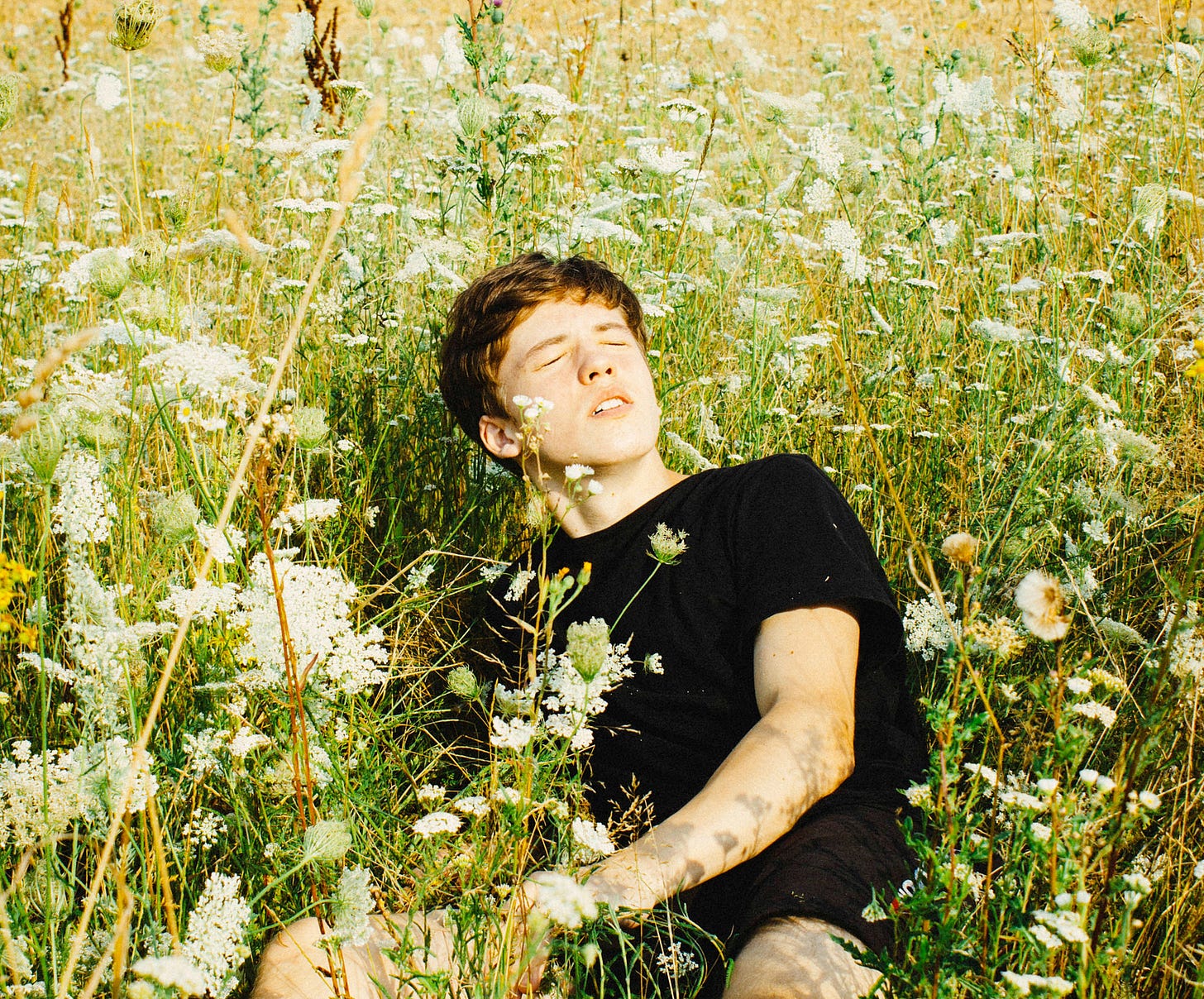 A young white man with short, slightly-ginger hair, wearing a black T-shirt and sitting and leaning back on his right arm in a field of tall green and amber grass and while flowers. It’s a sunny day, his eyes and closed and his face is turned towards the sun. A young white man with short, slightly-ginger hair, wearing a black T-shirt and sitting and leaning back on his right arm in a field of tall green and amber grass and while flowers. It’s a sunny day, his eyes and closed and his face is turned towards the sun.