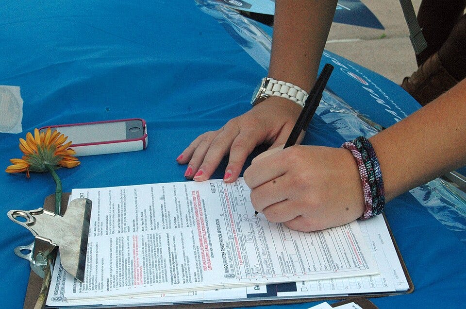 a closeup of a student's hands with nail polish and bracelets as they fill out a voter registration form on a clipboard on a blue tablecloth