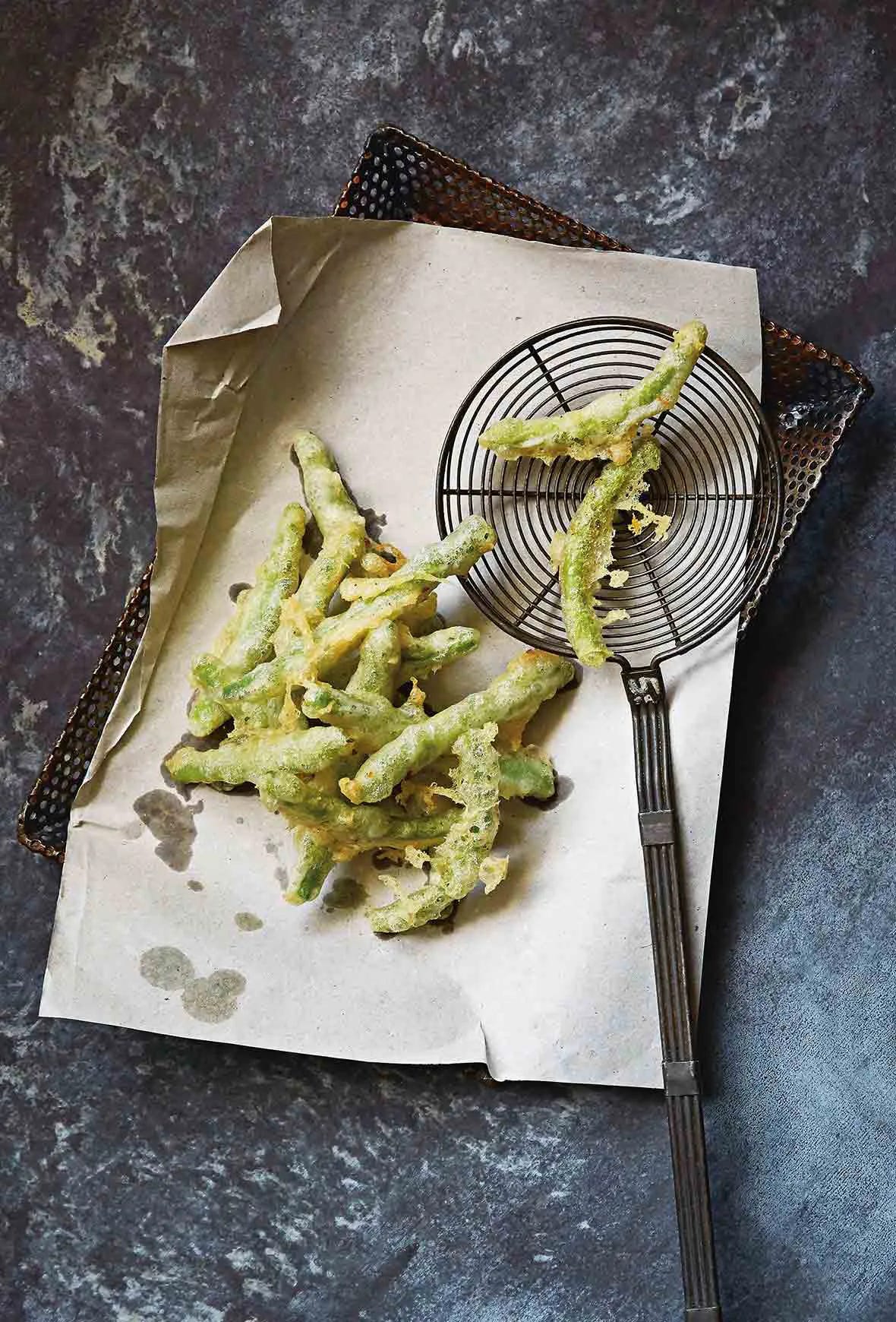A pile of Portuguese peixinhos da horta (tempura green beans) draining on brown paper in a strainer, a spider spatula nearby