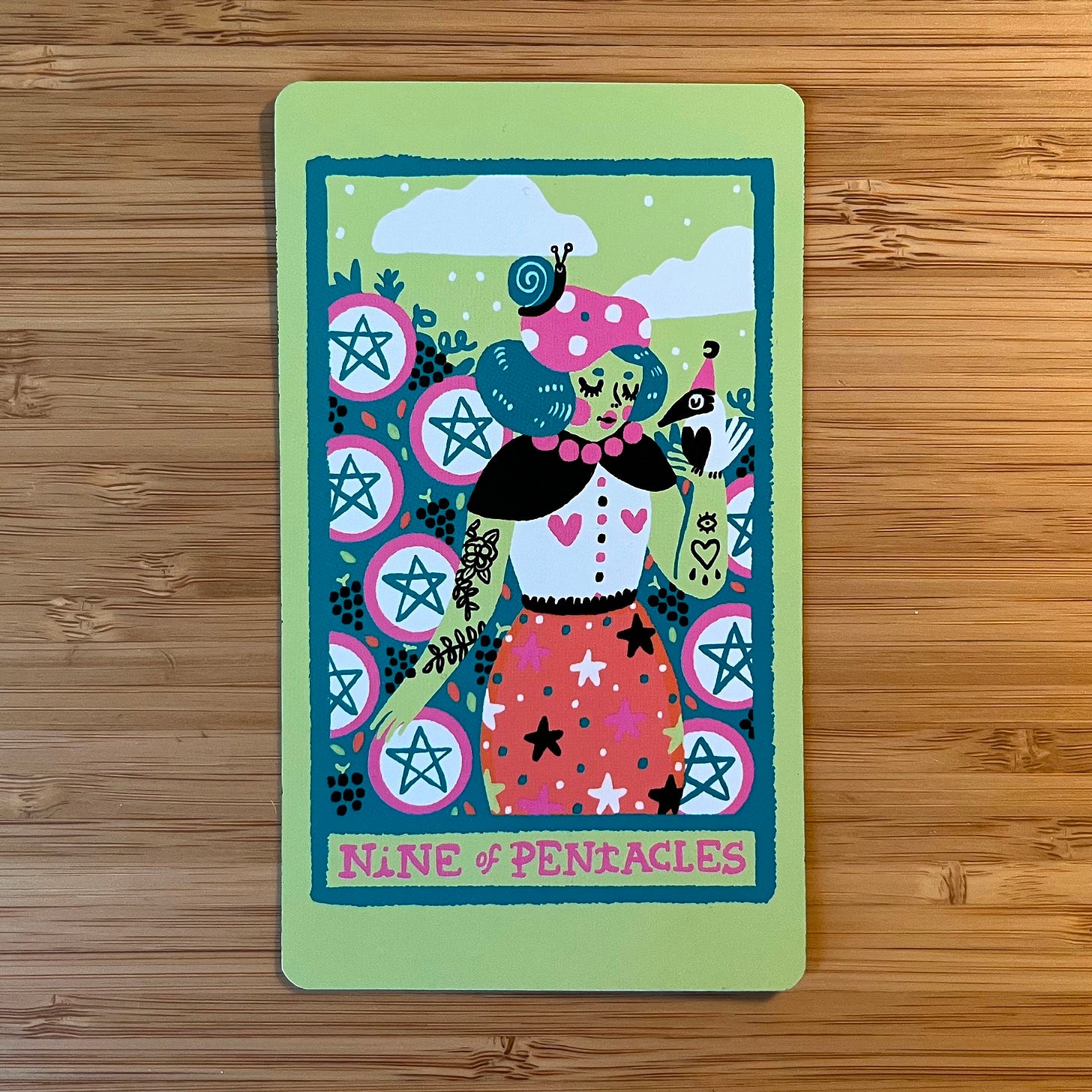 A pastel colored tarot card illustration of the Nine of Pentacles. The card depicts a figure in a whimsical outfit with a pink hat with white polka-dots, green hair, and a coral skirt with a pattern of pink, black, and white stars, and green and white polka dots. They have tattoos on both arms: one arm bears a vine with a single flower, the other arm bears the outline of a heart with an eye above it and three tear-shapes below. A bird is resting on their gloved left hand, and they stand in a lush garden of what looks like grape vines. The are surrounded by nine green, white, and pink pentacle symbols. The card background is green with white clouds in the sky, and the label at the bottom reads "Nine of Pentacles" in pink, capital letters. A pastel colored tarot card illustration of the Nine of Pentacles. The card depicts a figure in a whimsical outfit with a pink hat with white polka-dots, green hair, and a coral skirt with a pattern of pink, black, and white stars, and green and white polka dots. They have tattoos on both arms: one arm bears a vine with a single flower, the other arm bears the outline of a heart with an eye above it and three tear-shapes below. A bird is resting on their gloved left hand, and they stand in a lush garden of what looks like grape vines. The are surrounded by nine green, white, and pink pentacle symbols. The card background is green with white clouds in the sky, and the label at the bottom reads "Nine of Pentacles" in pink, capital letters.