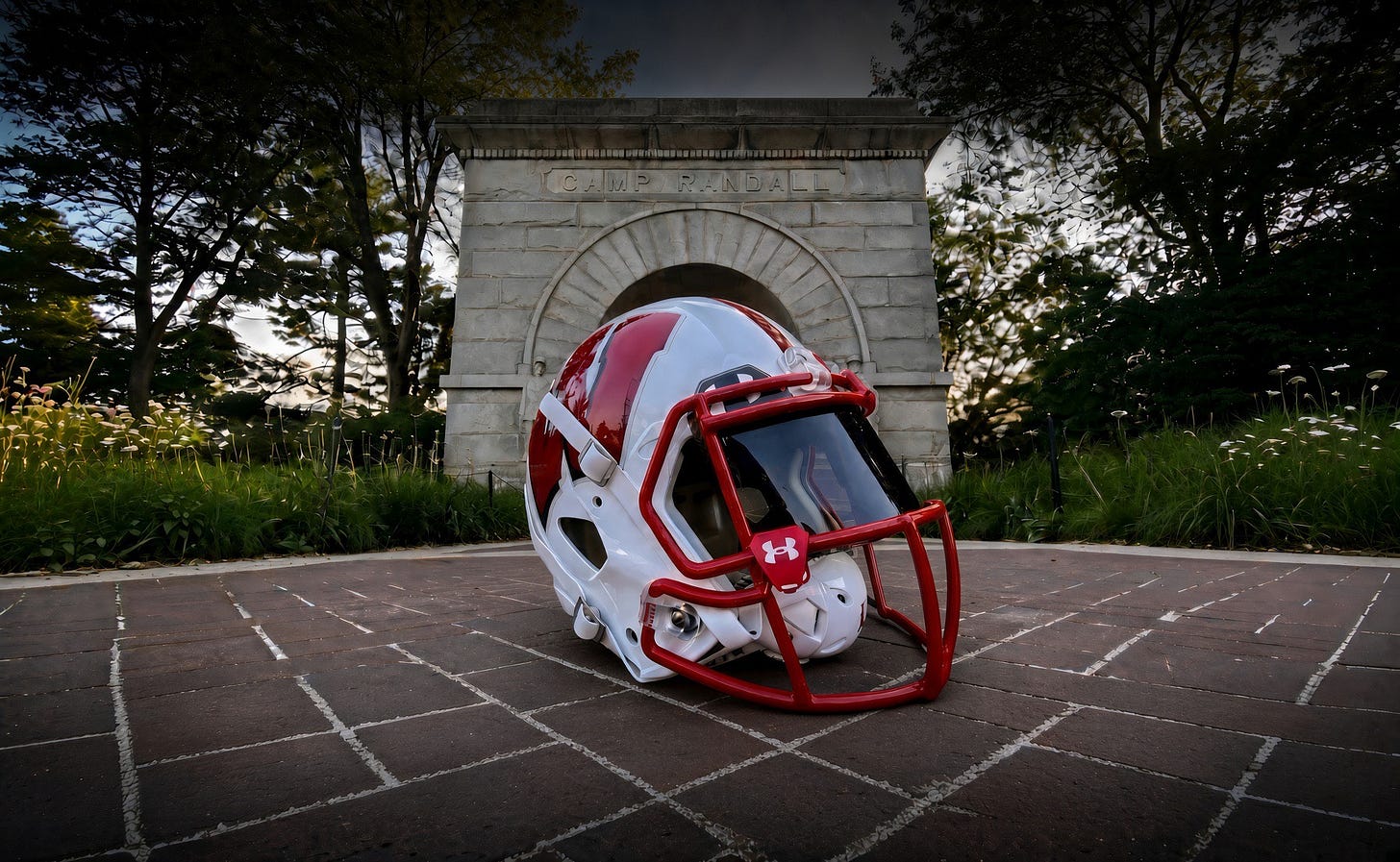 A red and white Wisconsin Badgers football helmet with a red facemask sits on a brick walkway in front of the Camp Randall Memorial Arch.