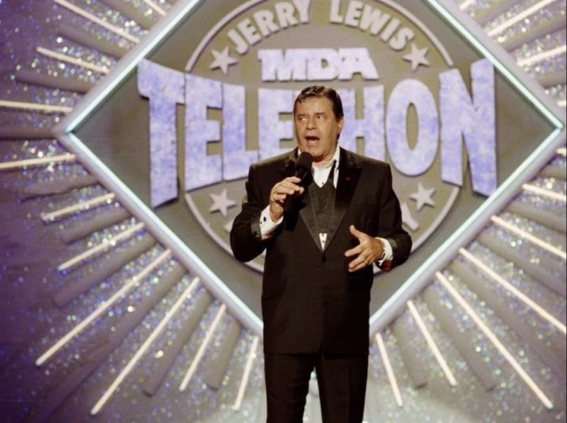 : Dressed in a dark suit, middle aged Jerry Lewis holds a microphone toward his open mouth. He gestures with his left hand. He stands before a banner reading MDA telethon.)