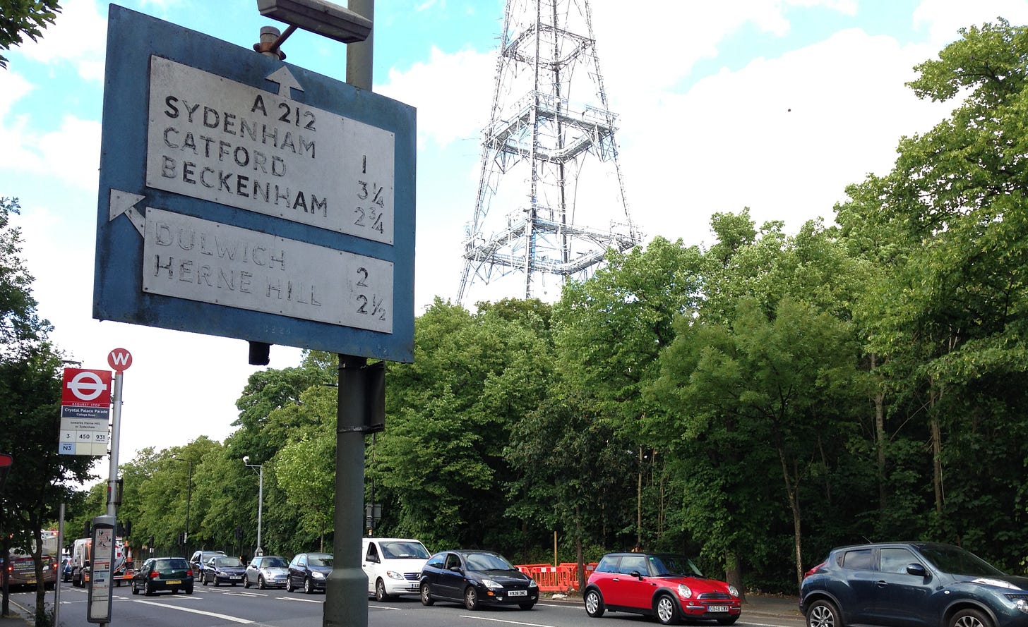 Photograph of a faded blue and white direction sign in front of a busy street. A transmitter mast is visible above the trees in the background Photograph of a faded blue and white direction sign in front of a busy street. A transmitter mast is visible above the trees in the background
