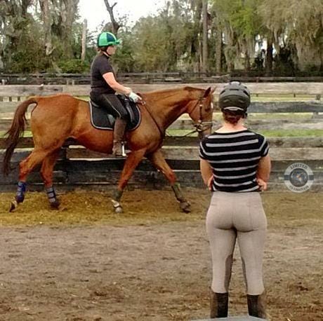 A young woman stands in an arena watching someone riding a horse nearby