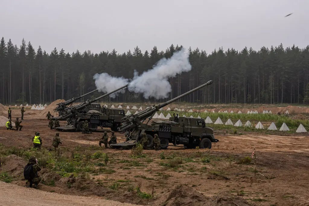 Self-propelled howitzers fire into the air in a field surrounded by woods. A handful of reservists stand or kneel nearby.