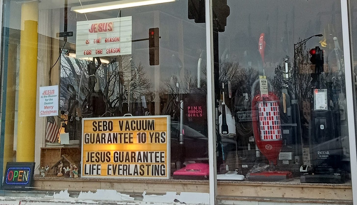 Shopfront window of a vacuum store. Signs in the window read "Open", "JESUS is the reason for the Season Merry CHRISTmas", "JESUS is the reason for the season", and "Sebo Vacuum guarantee 10 years. Jesus guarantee life everlasting"