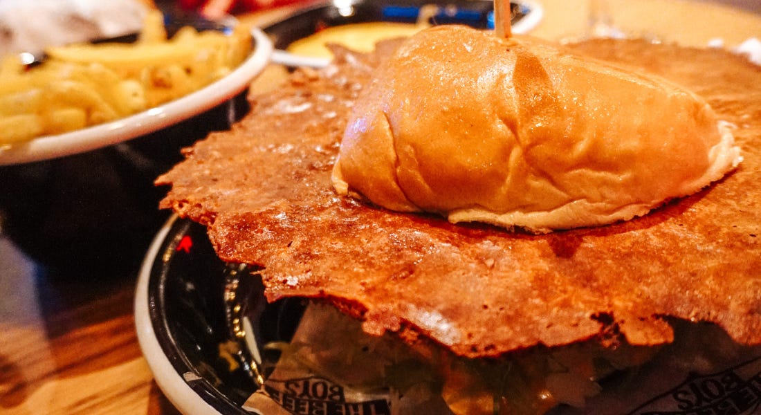 A burger with a cheese skirt in the middle on a black plate, with a plate of fries above it